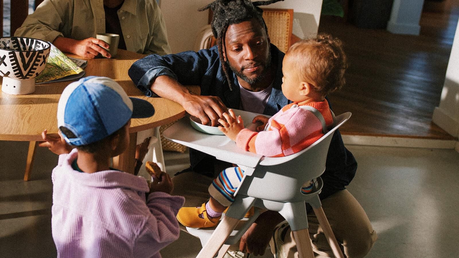 A man interacts with a baby in a high chair at a table, another child nearby.