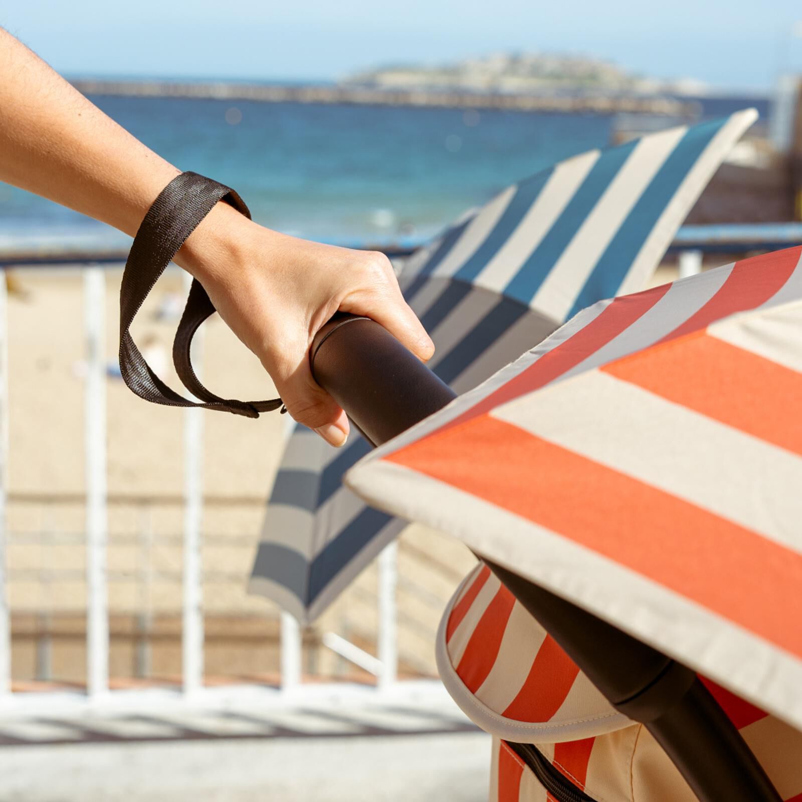 Main tenant une poussette STOKKE YOYO avec auvent rouge et blanc au bord de la mer.