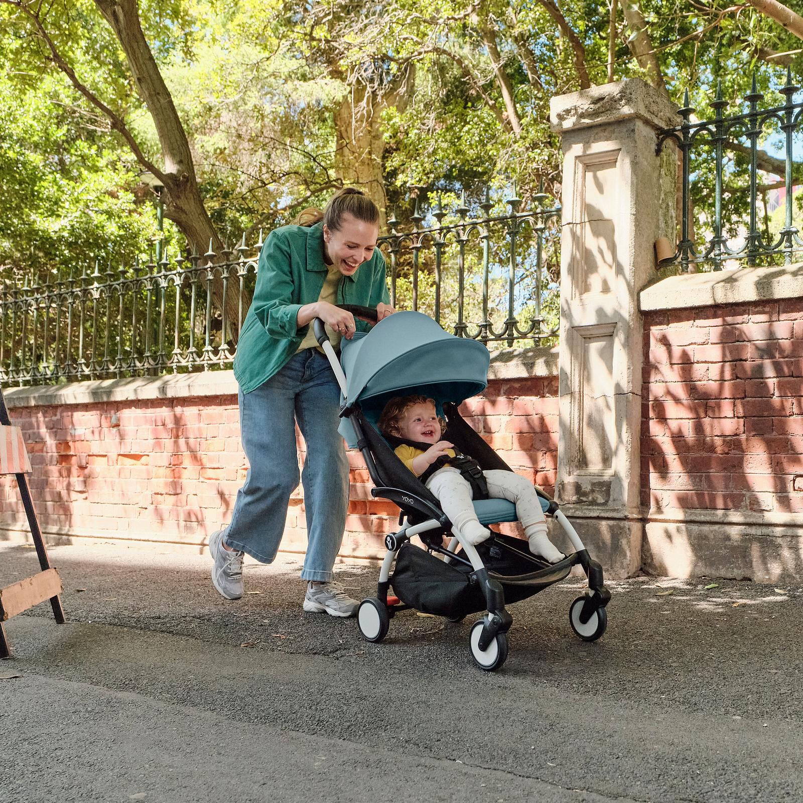 Eine Frau schiebt ein glückliches Baby in einem blauen STOKKE YOYO Kinderwagen auf einem sonnigen Weg.