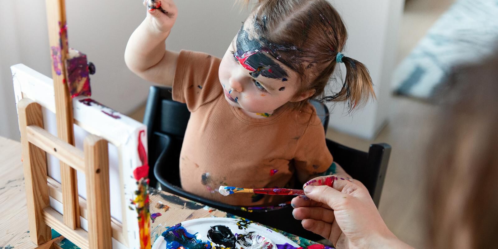 Paint-covered toddler in a black Tripp Trapp high chair reaches for a paintbrush.