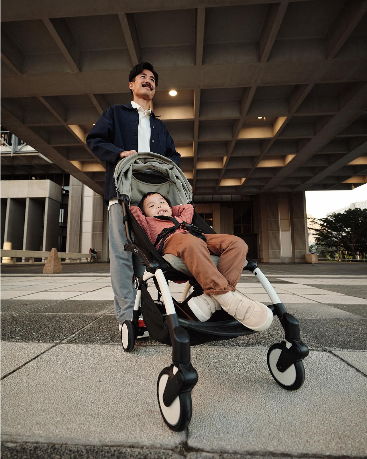 Smiling man pushes a happy child in a STOKKE YOYO stroller under a concrete building.