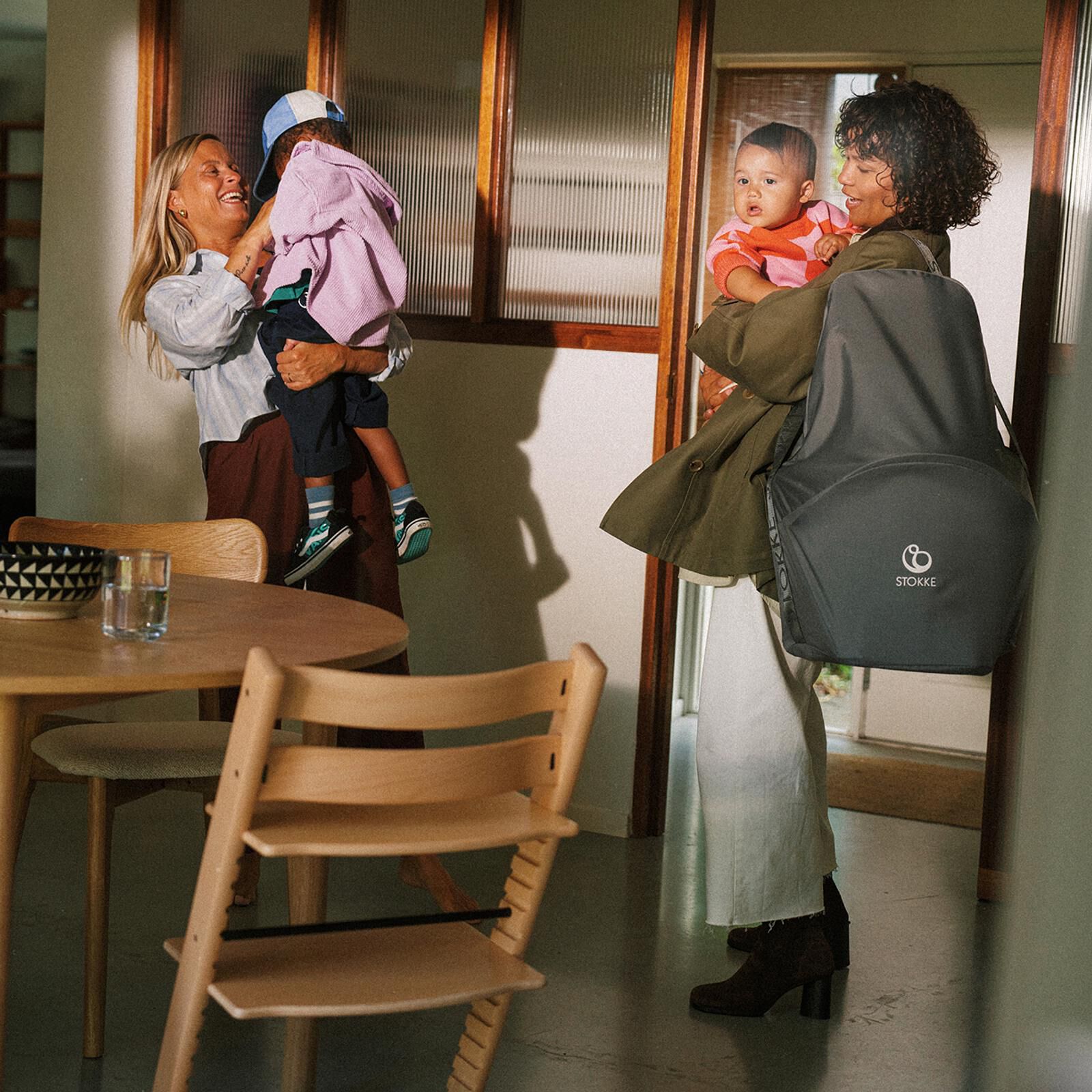 Dos mujeres abrazan a niños; una lleva un cochecito STOKKE YOYO. Una silla Tripp Trapp está en primer plano.