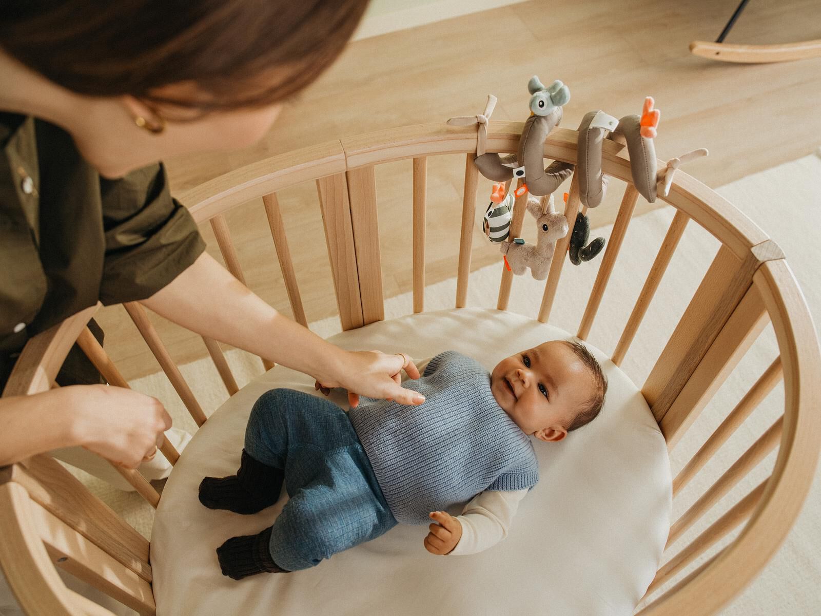 En baby i en oval trebakhylle smiler opp mot en foreldres hånd, med en uro over seg.