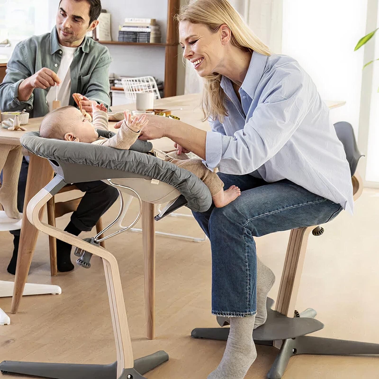Smiling parents interact with their baby in a grey newborn high chair at a wooden table.