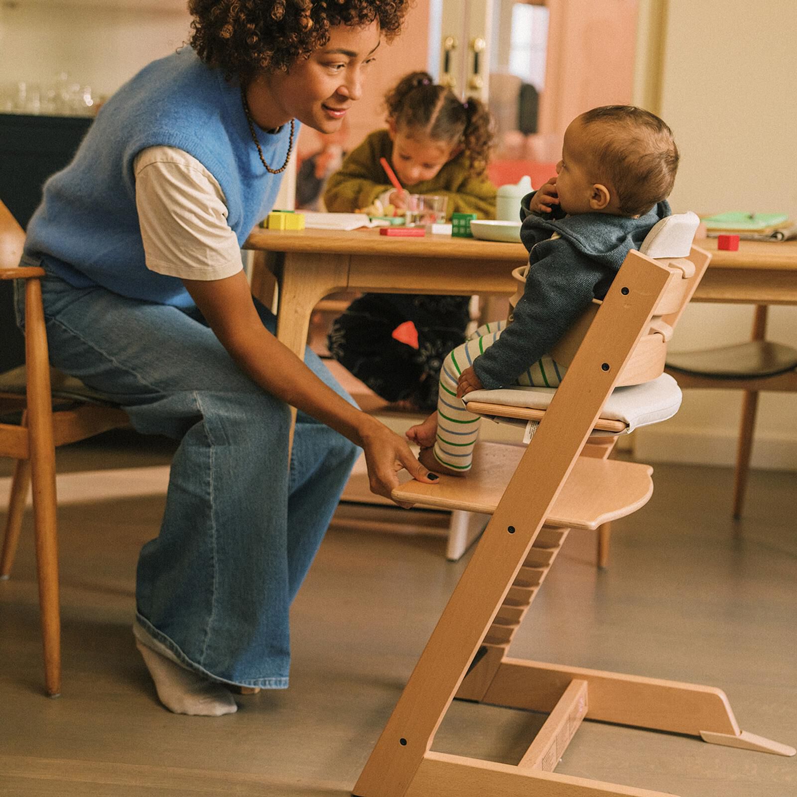 A woman adjusts a Tripp Trapp high chair for a baby, with another child in the background.