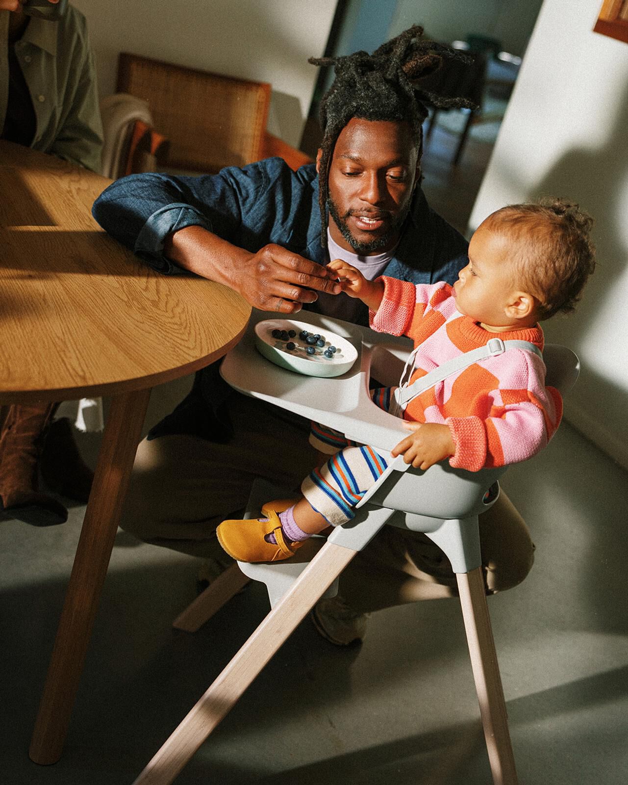 A man feeds blueberries to a baby in a Tripp Trapp high chair at a wooden table.