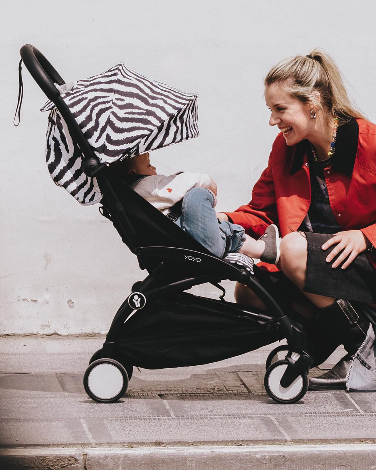 Una mujer sonriente se arrodilla junto a un cochecito STOKKE YOYO con un bebé bajo un dosel con estampado de cebra.