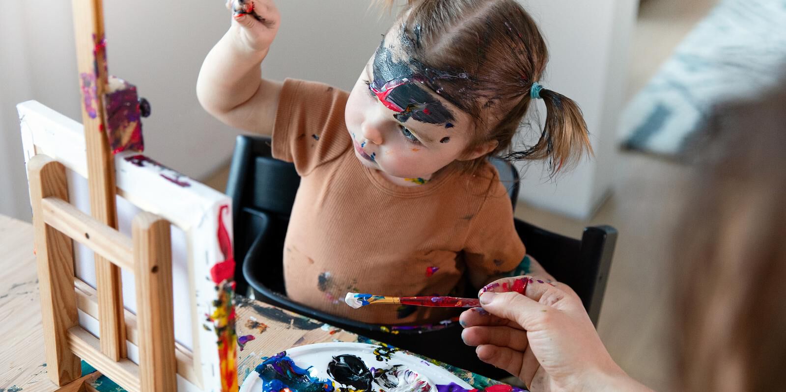 Paint-covered toddler in a black Tripp Trapp high chair reaches for a paintbrush.