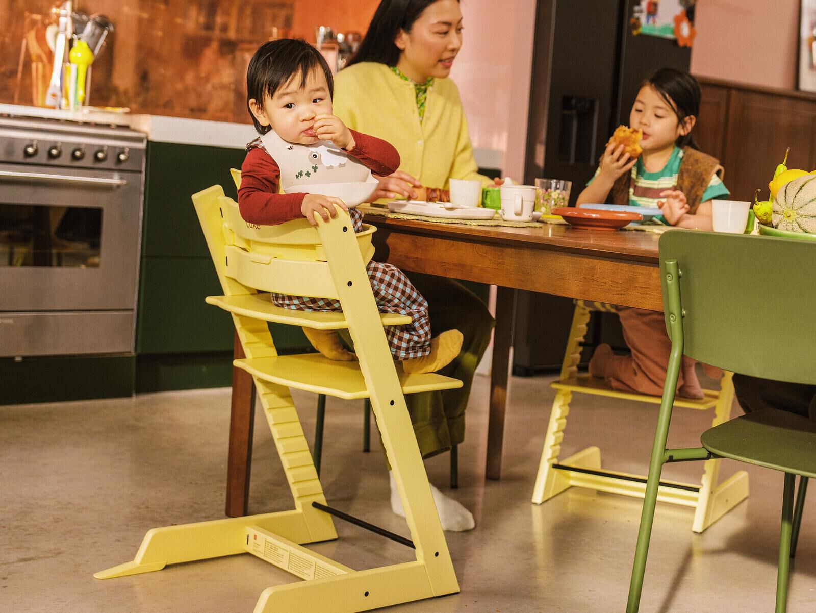 Et lite barn spiser i en gul Tripp Trapp barnestol, sammen med familien ved et kjøkkenbord.