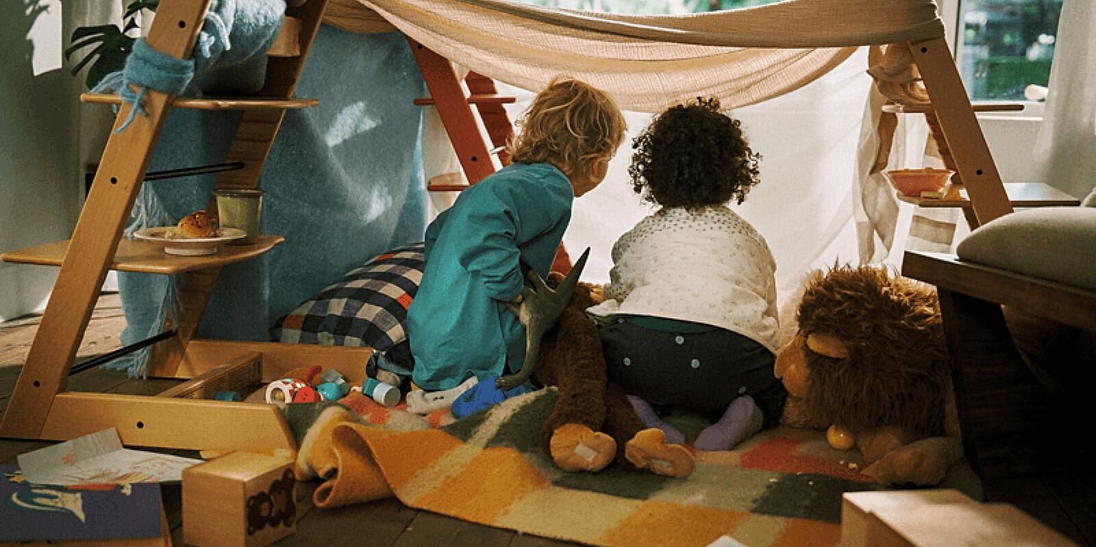 Two children play in a cozy blanket fort built around a Tripp Trapp chair.