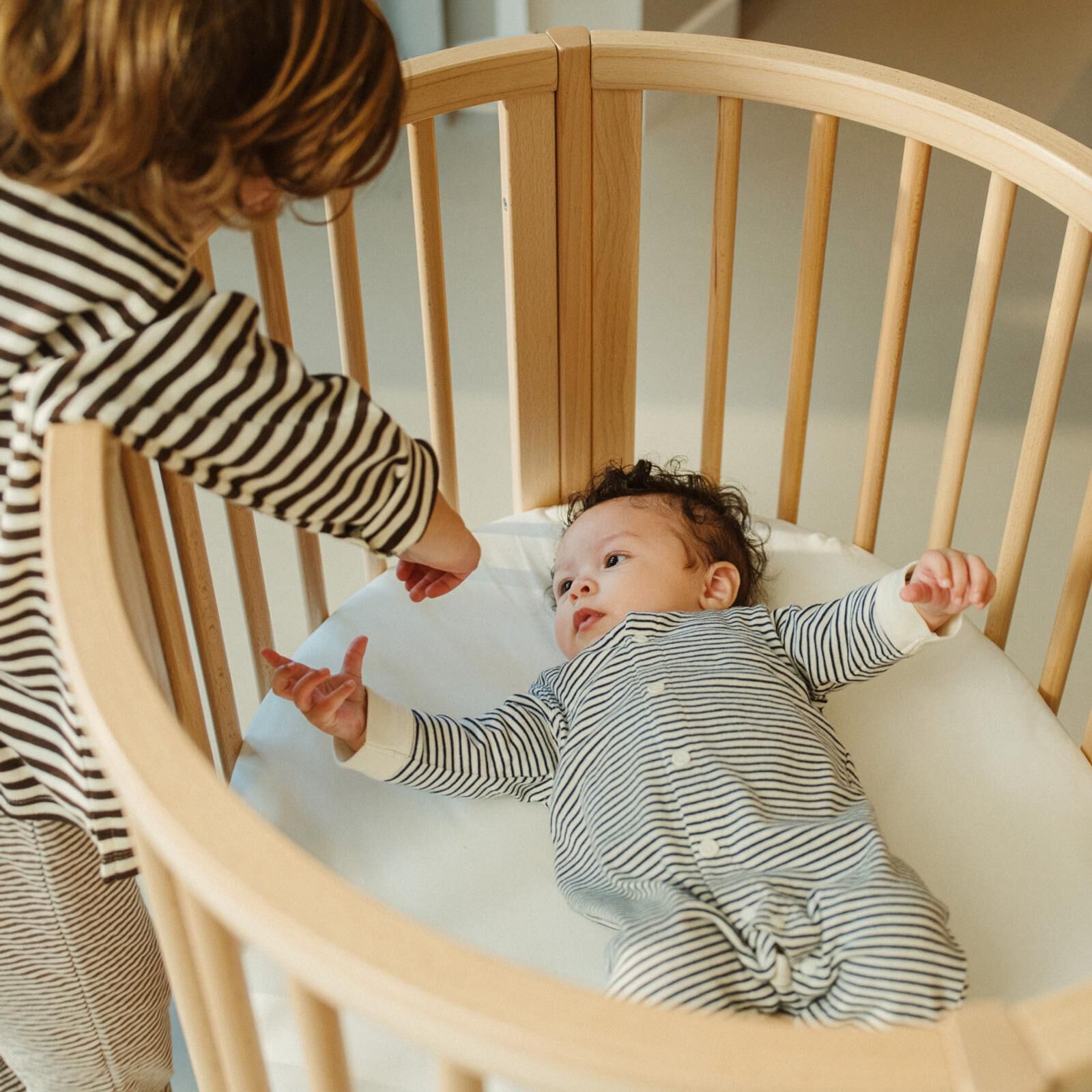An older child leans over a wooden crib, reaching toward a baby in a striped onesie.