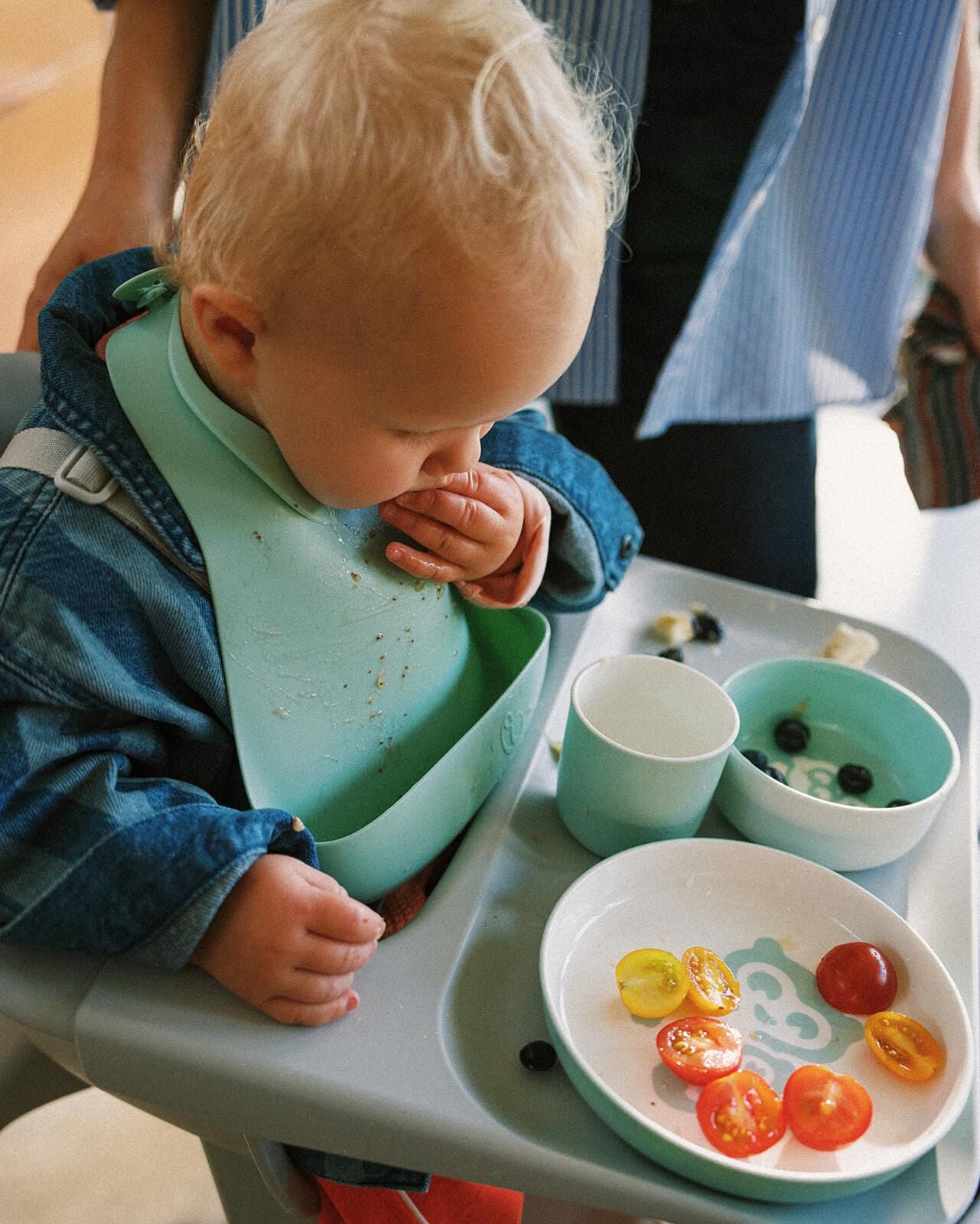 Blonde baby in a grey high chair, wearing a denim jacket and mint bib, eating cherry tomatoes and blueberries.