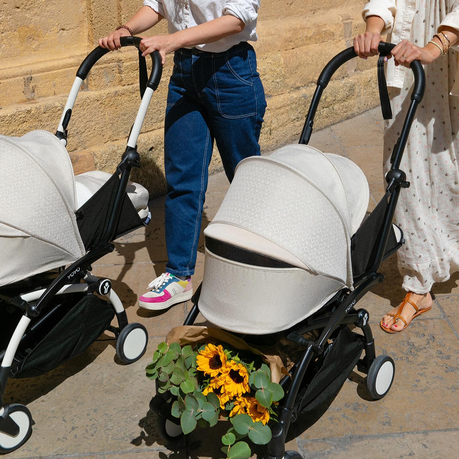Two people push two beige STOKKE YOYO strollers. One has sunflowers in its basket.