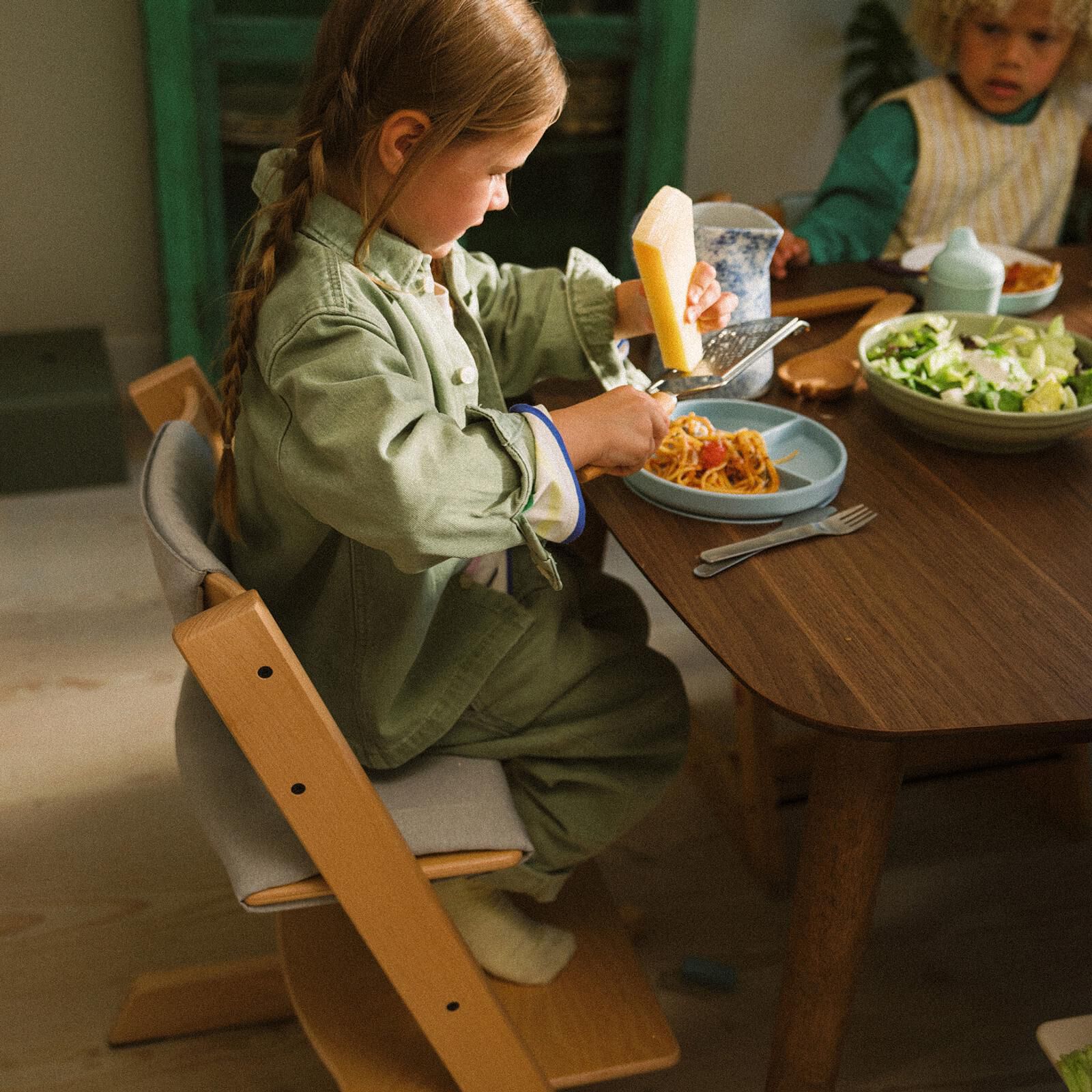 A child grates cheese onto pasta at a table, sitting in a Tripp Trapp chair.