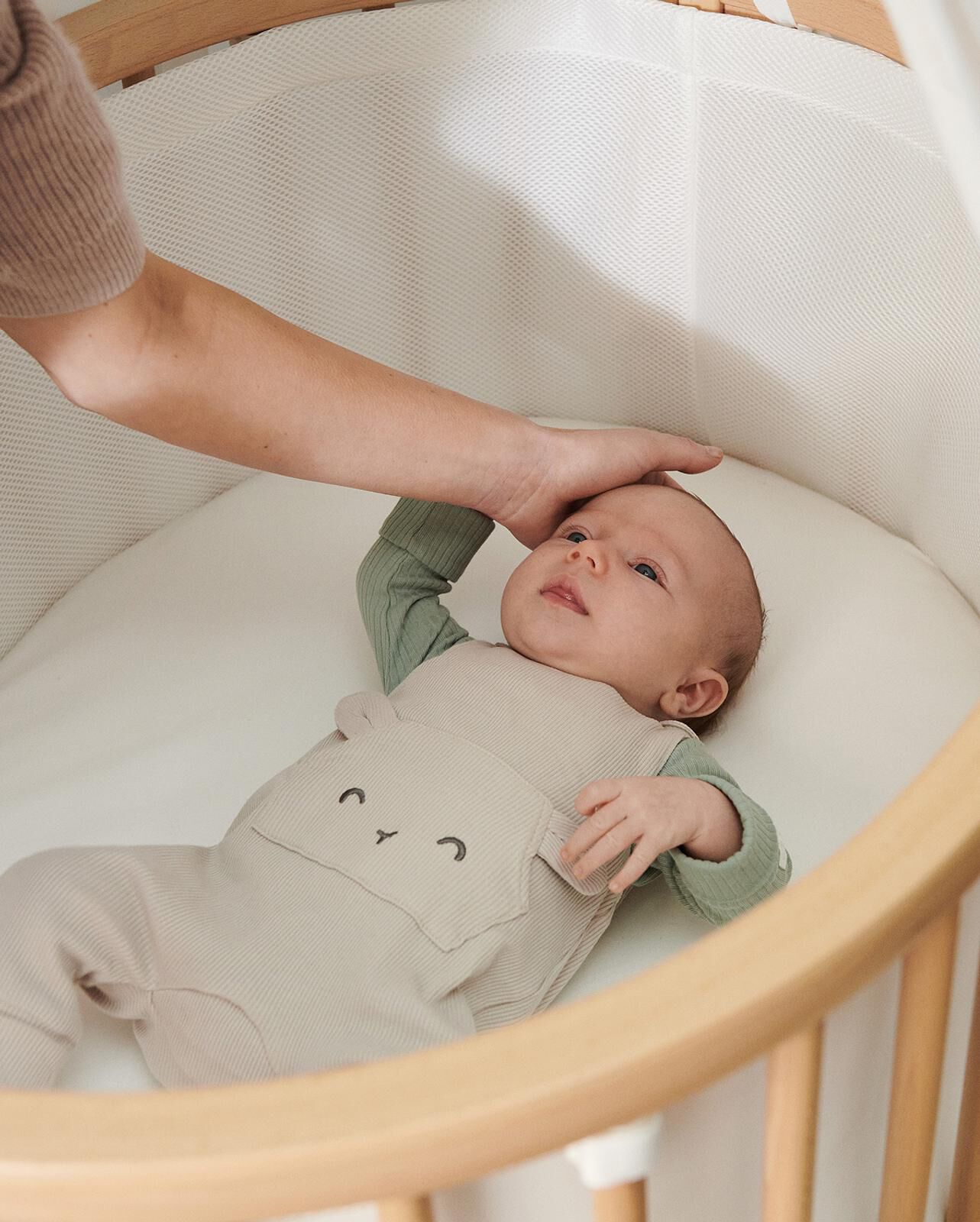 Un bébé dans un berceau ovale regarde vers le haut tandis que la main d'un adulte touche doucement sa tête.