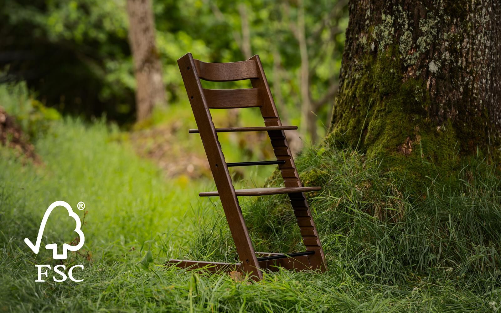 A brown Tripp Trapp chair stands in a forest clearing next to a mossy tree trunk. FSC logo.