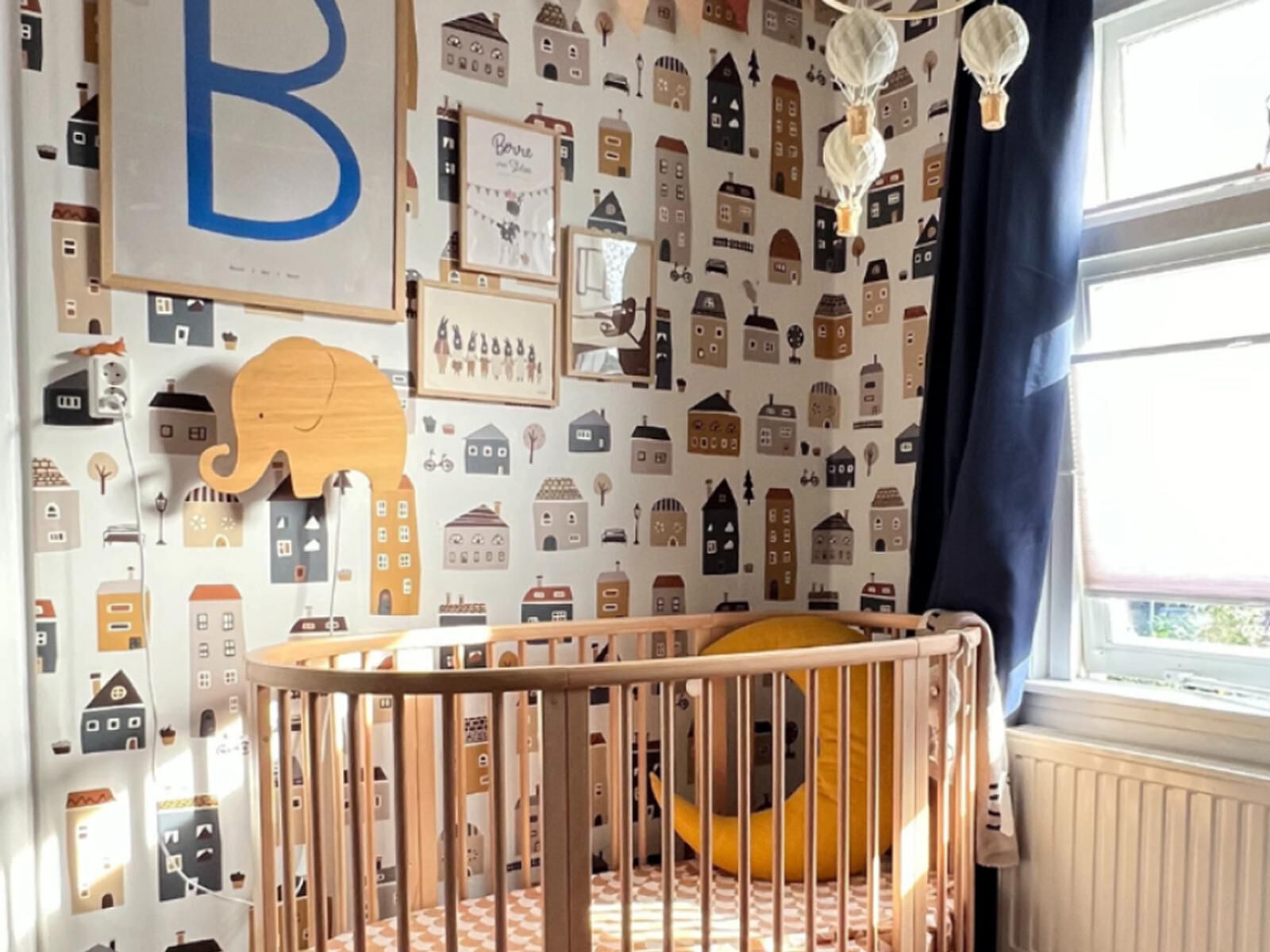 Une chambre de bébé confortable avec un berceau ovale en bois, du papier peint maison et une applique murale éléphant en bois.