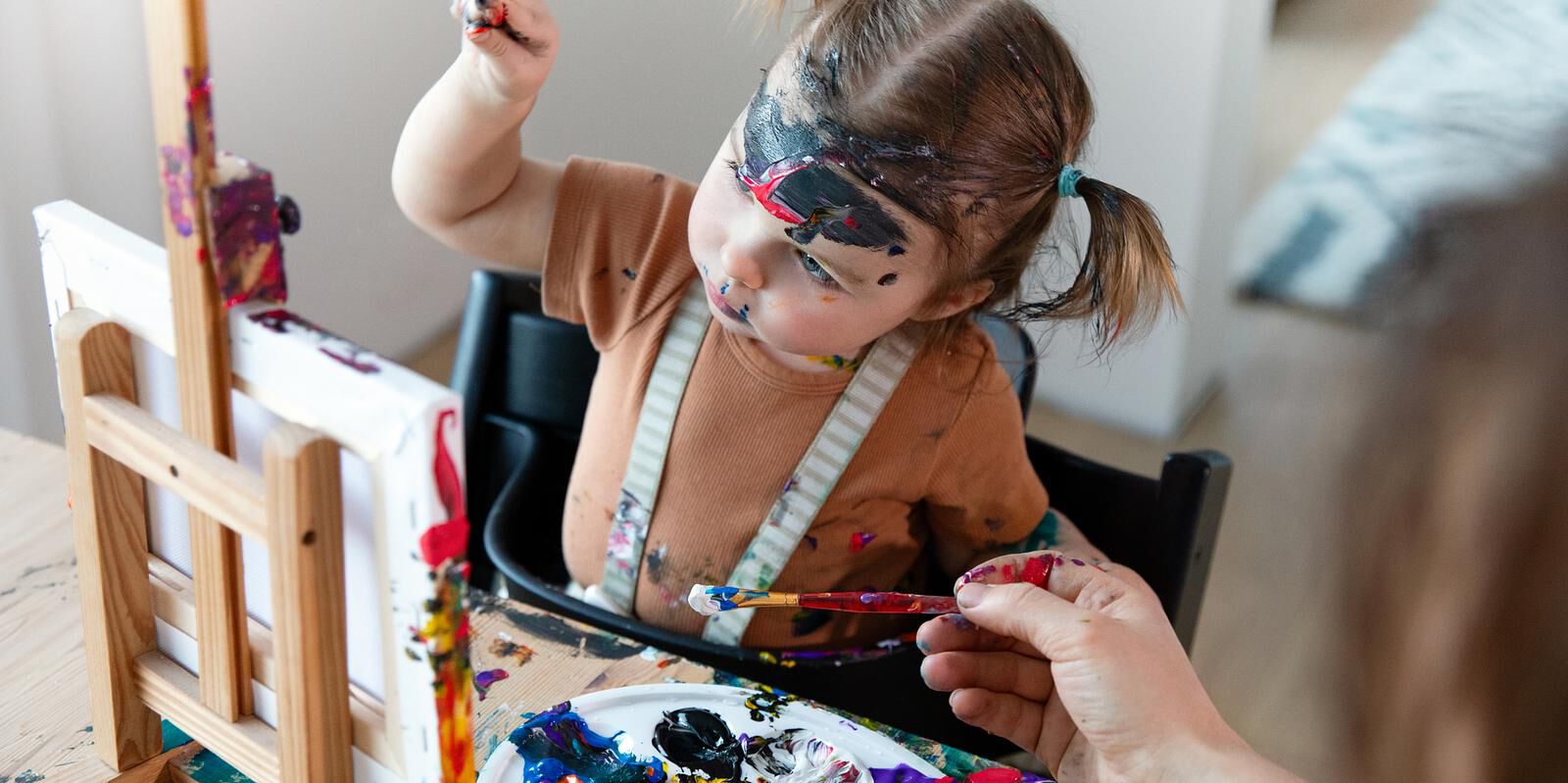 A messy toddler in a Tripp Trapp high chair paints at an easel, face covered in paint.