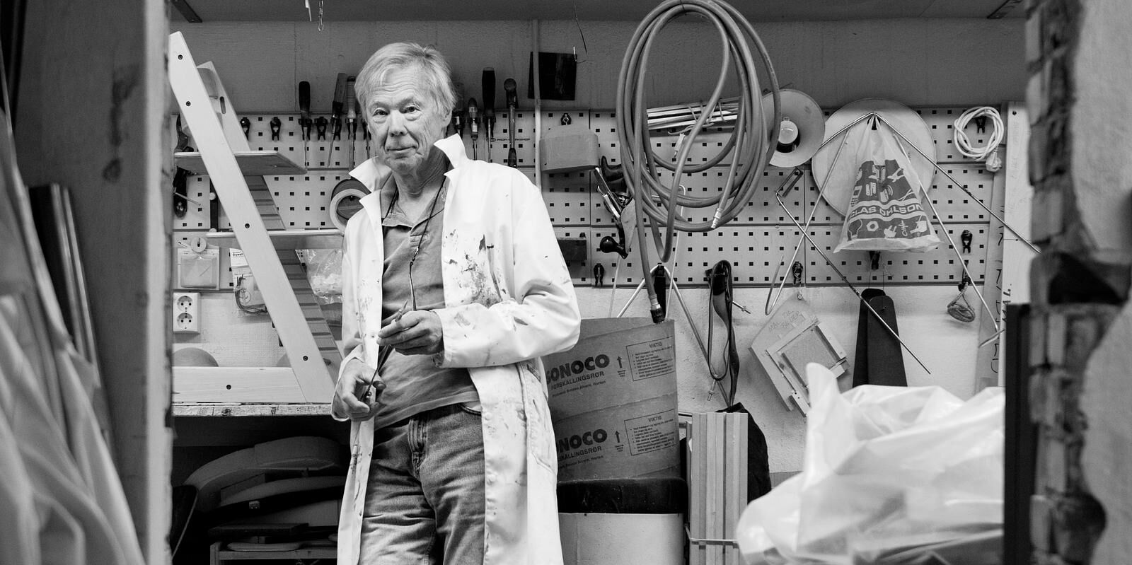 Elderly man in a paint-stained lab coat holds a tool in a cluttered workshop with a Tripp Trapp chair.