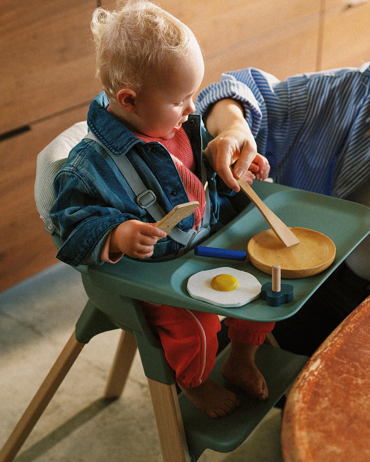 Un bébé joue avec de la nourriture factice sur un plateau dans une chaise haute verte, avec la main d'un adulte qui l'aide.