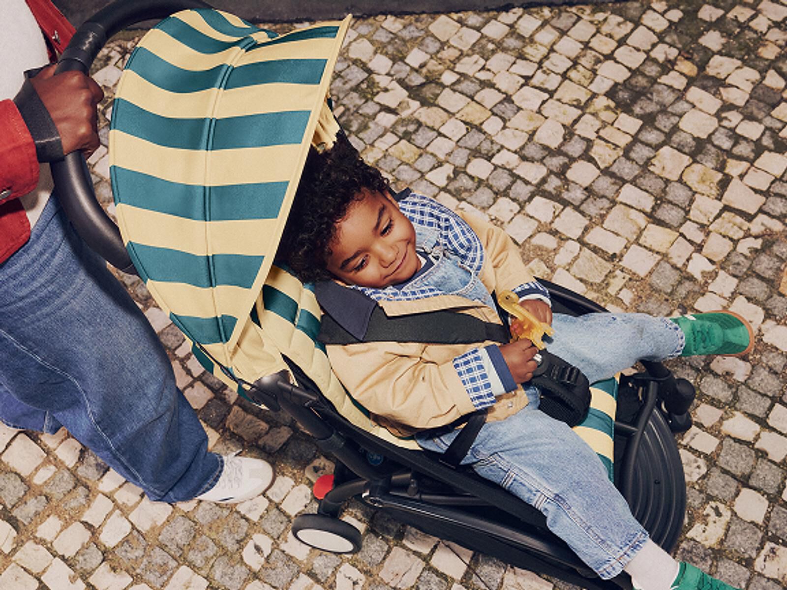 A smiling toddler in a STOKKE YOYO stroller with a striped canopy on a cobblestone path.