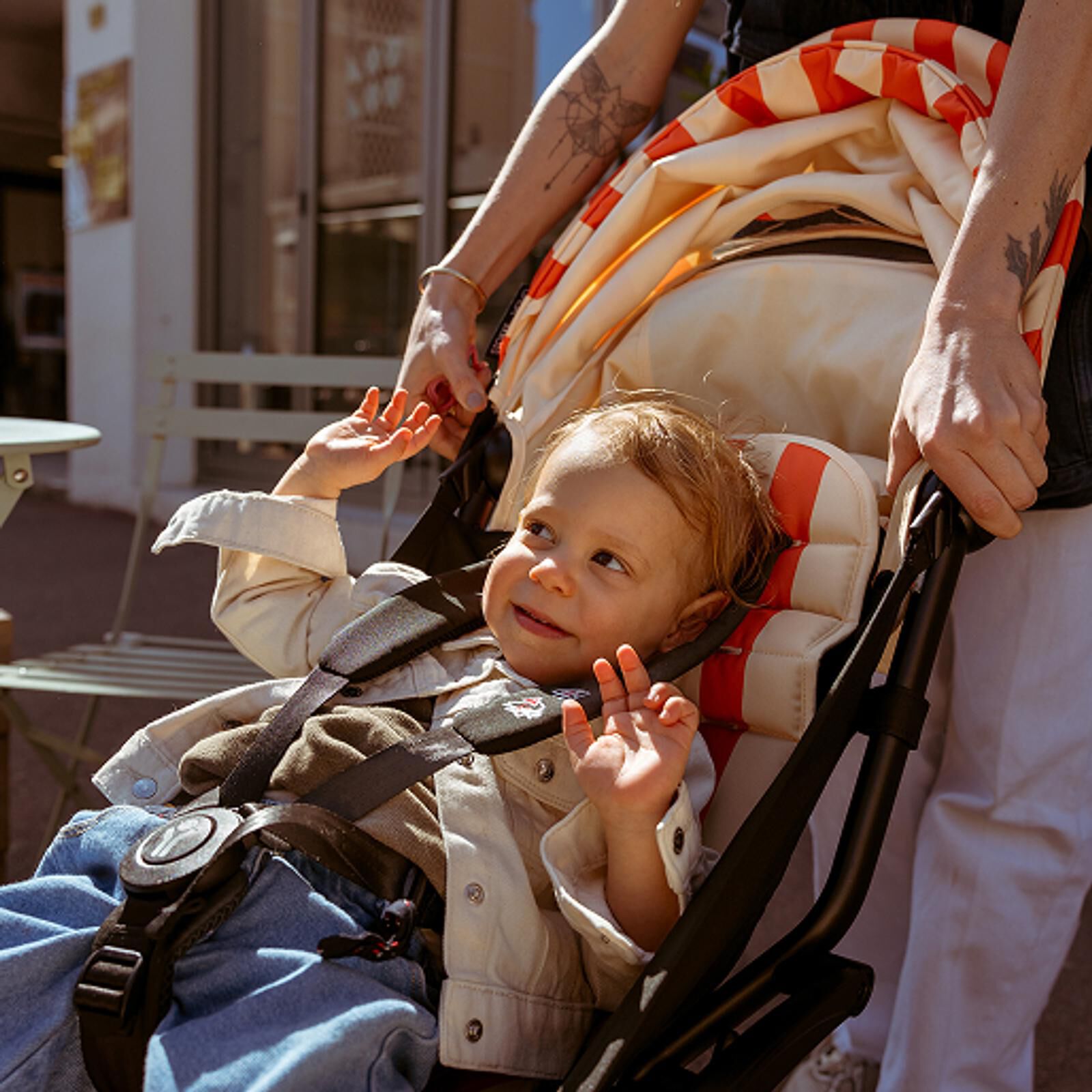 Glückliches Kind lächelt, Hände erhoben, in einem STOKKE YOYO Kinderwagen, der von einem Erwachsenen im Freien geschoben wird.