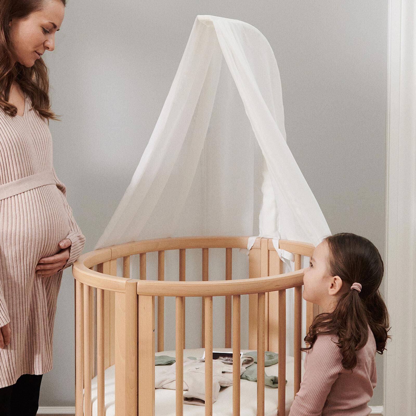Une femme enceinte et une fille regardent un berceau en bois avec un baldaquin, rempli de vêtements de bébé.