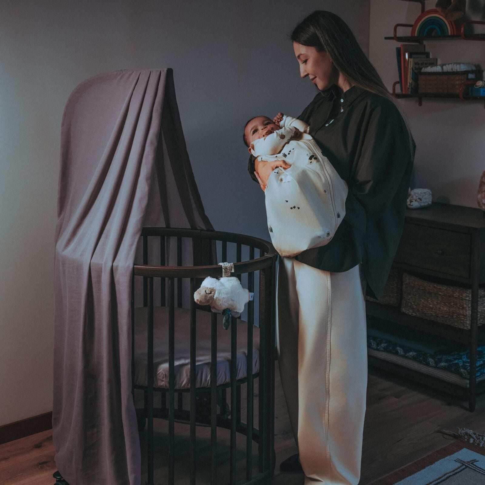 Vrouw houdt baby in slaapzak, naast een rond wiegje met hemel.