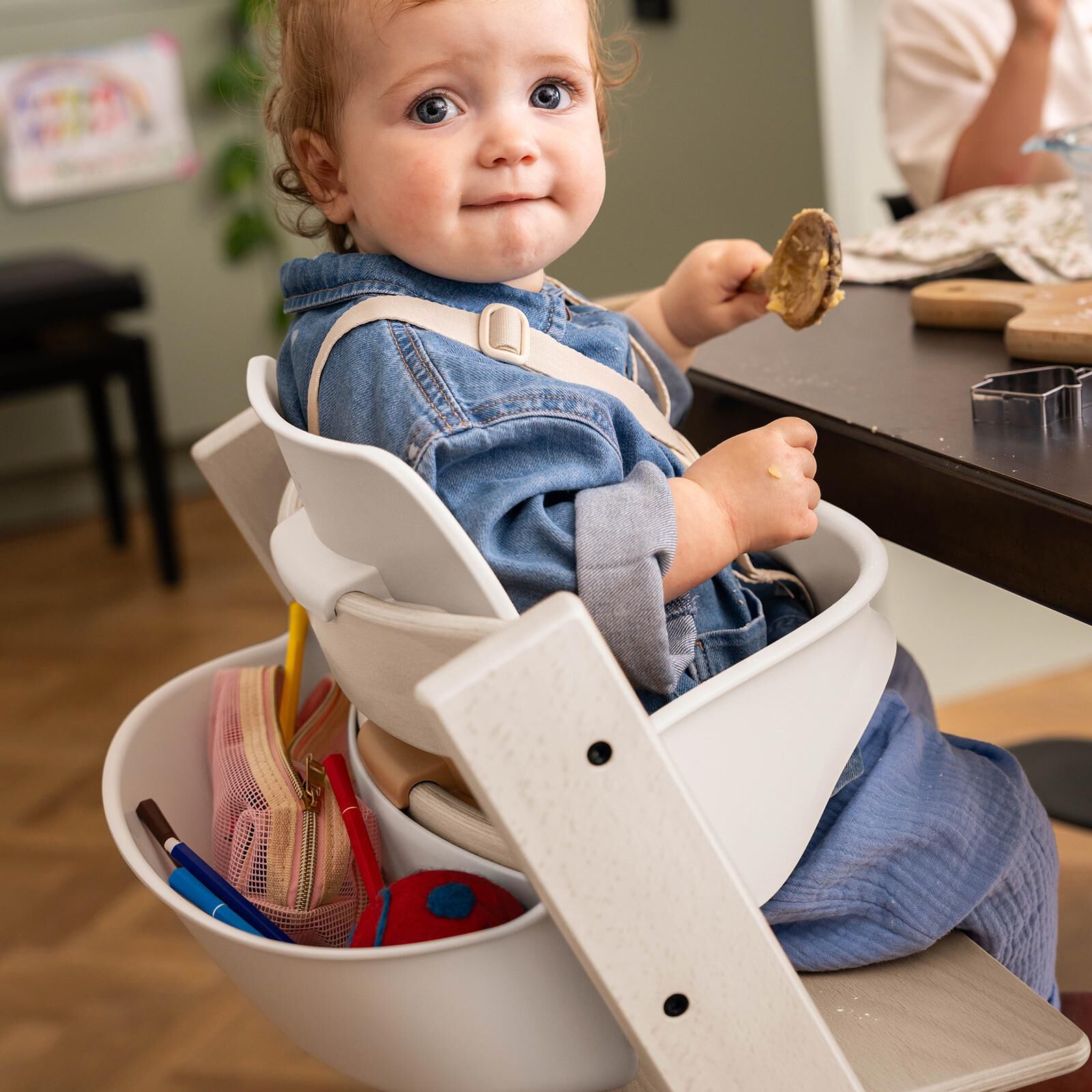 A baby sits in a white Tripp Trapp high chair, holding a spoon, with art supplies on the side.