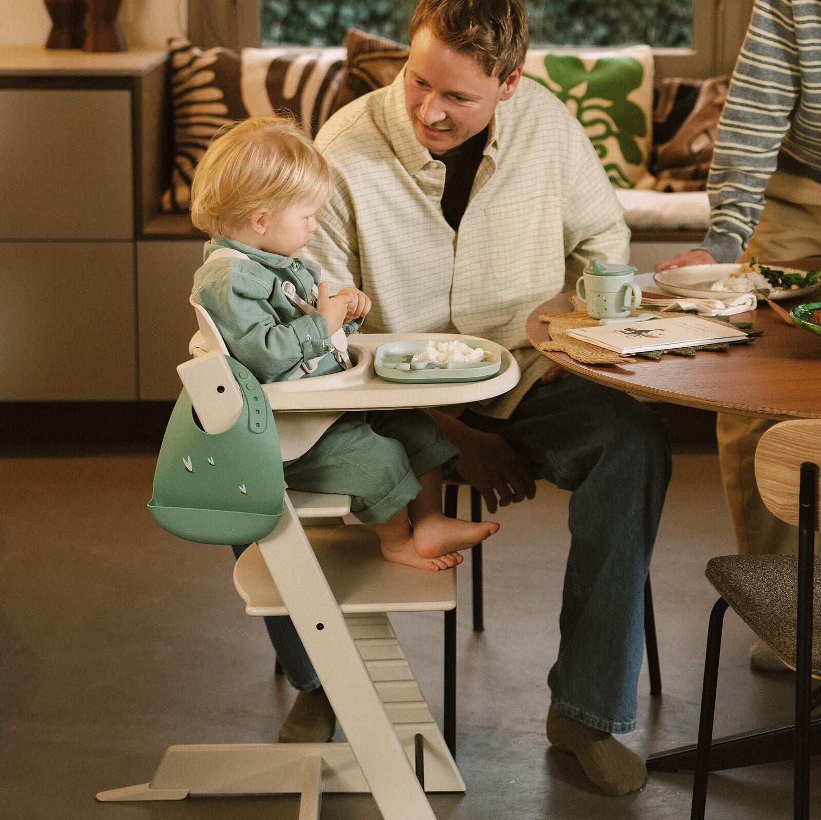 A man smiles at a child in a green bib eating in a white Tripp Trapp high chair.