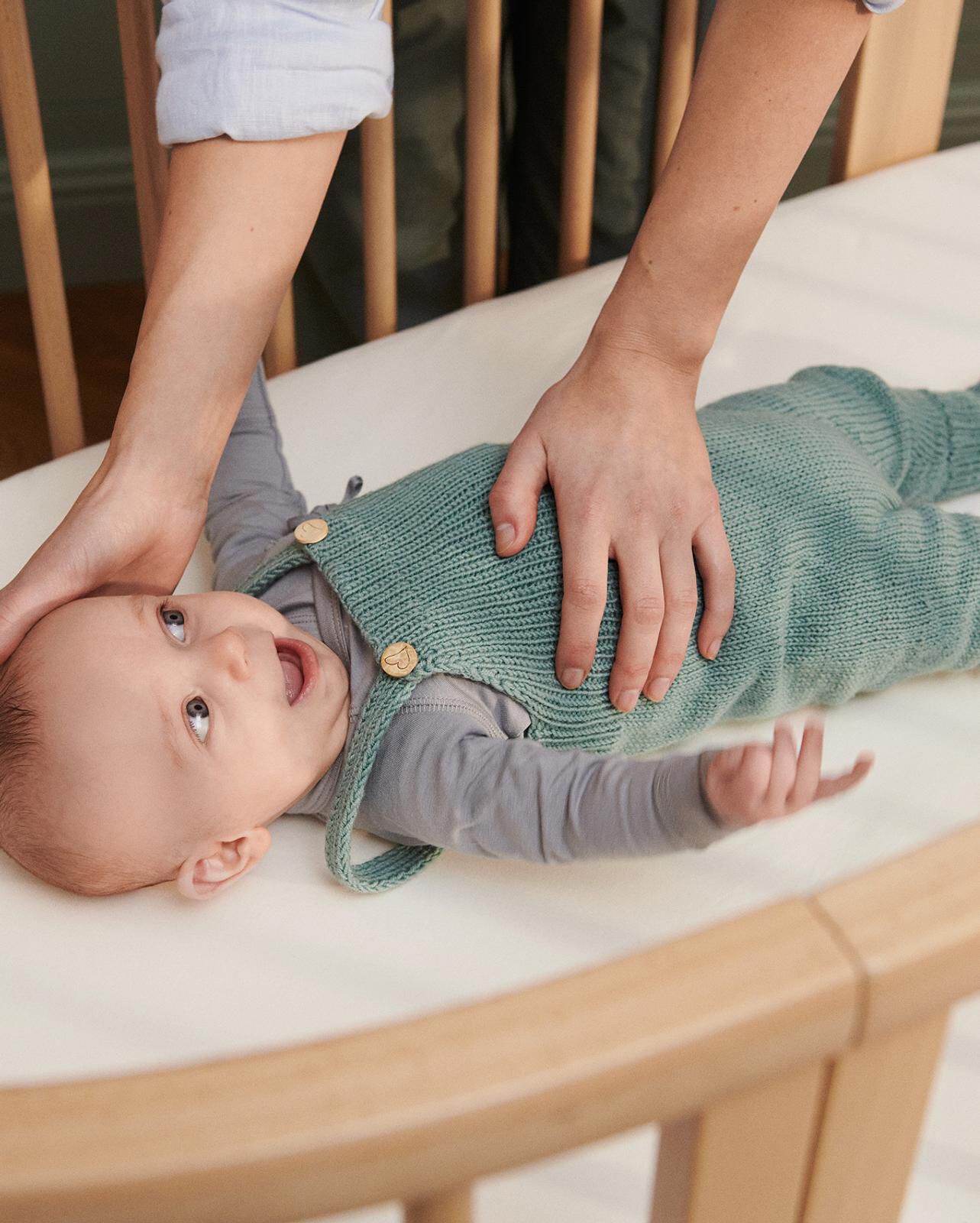 Adult hands gently tend to a happy baby in a wooden crib, wearing a teal knitted outfit.