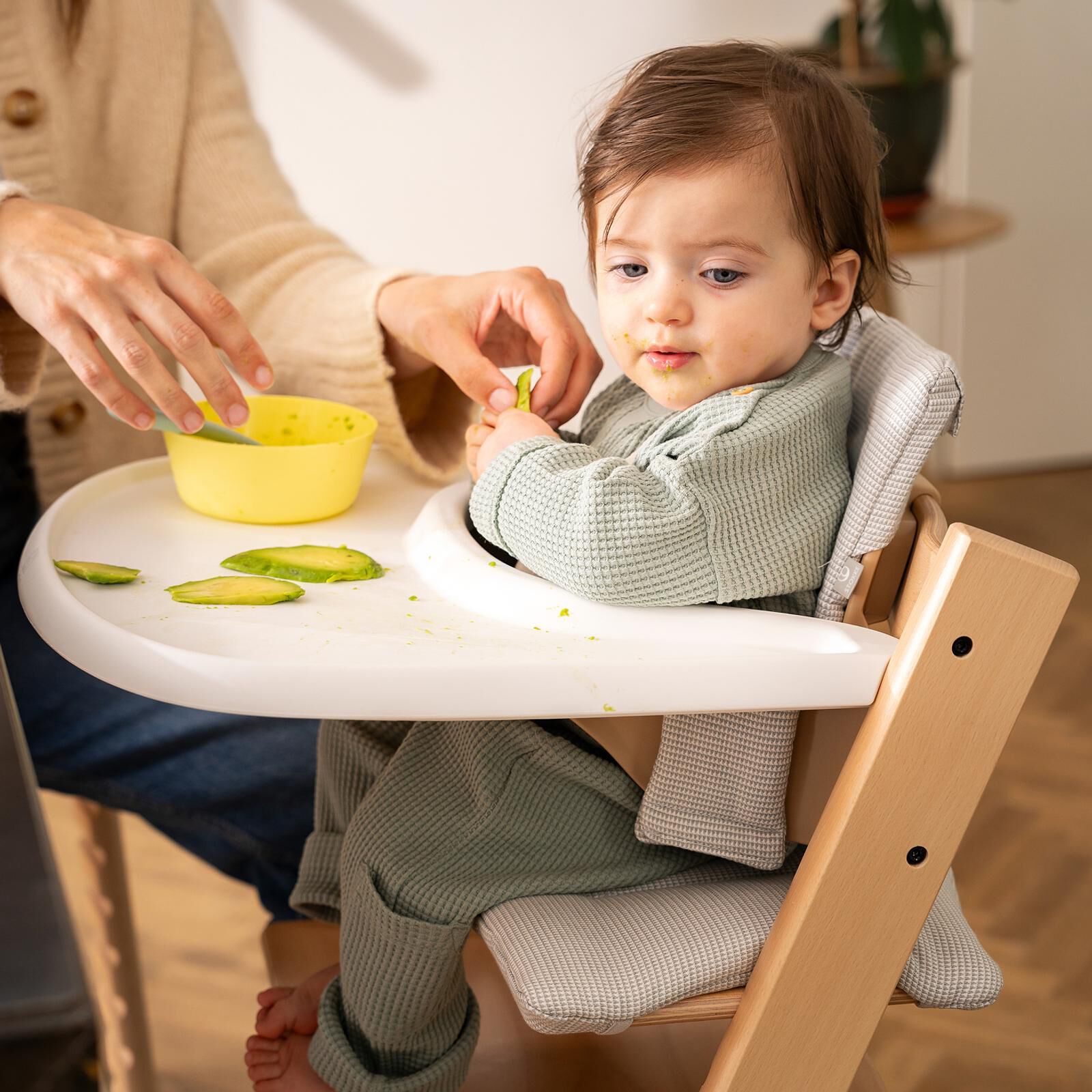 A baby with avocado on their face sits in a Tripp Trapp high chair, eating avocado.