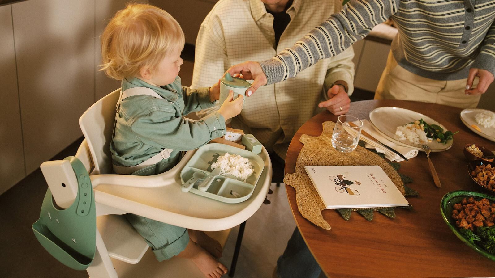 Een kind in een kinderstoel reikt naar een tuitbeker die een volwassene aanreikt aan een eettafel.