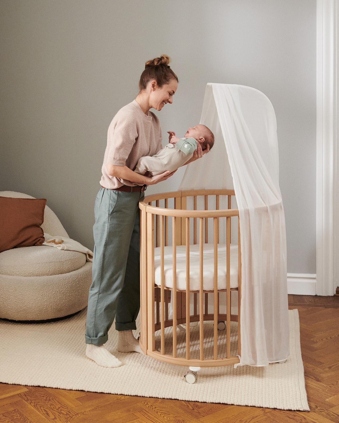 A smiling woman holds a baby over a round wooden crib with a white canopy.