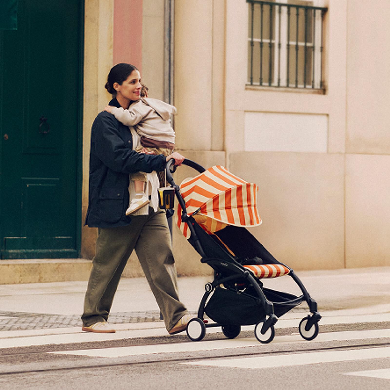 Frau trägt ein Kind und schiebt einen STOKKE YOYO Kinderwagen mit orange-weiß gestreiftem Verdeck auf einer Straße.