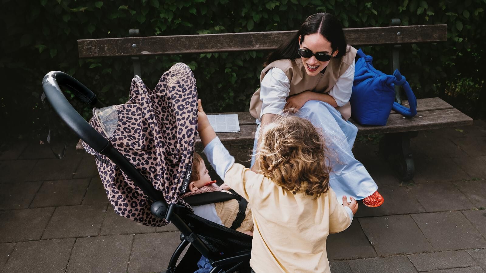 Une femme souriante assise sur un banc regarde un enfant tendre la main vers un bébé dans une poussette STOKKE YOYO à imprimé léopard.