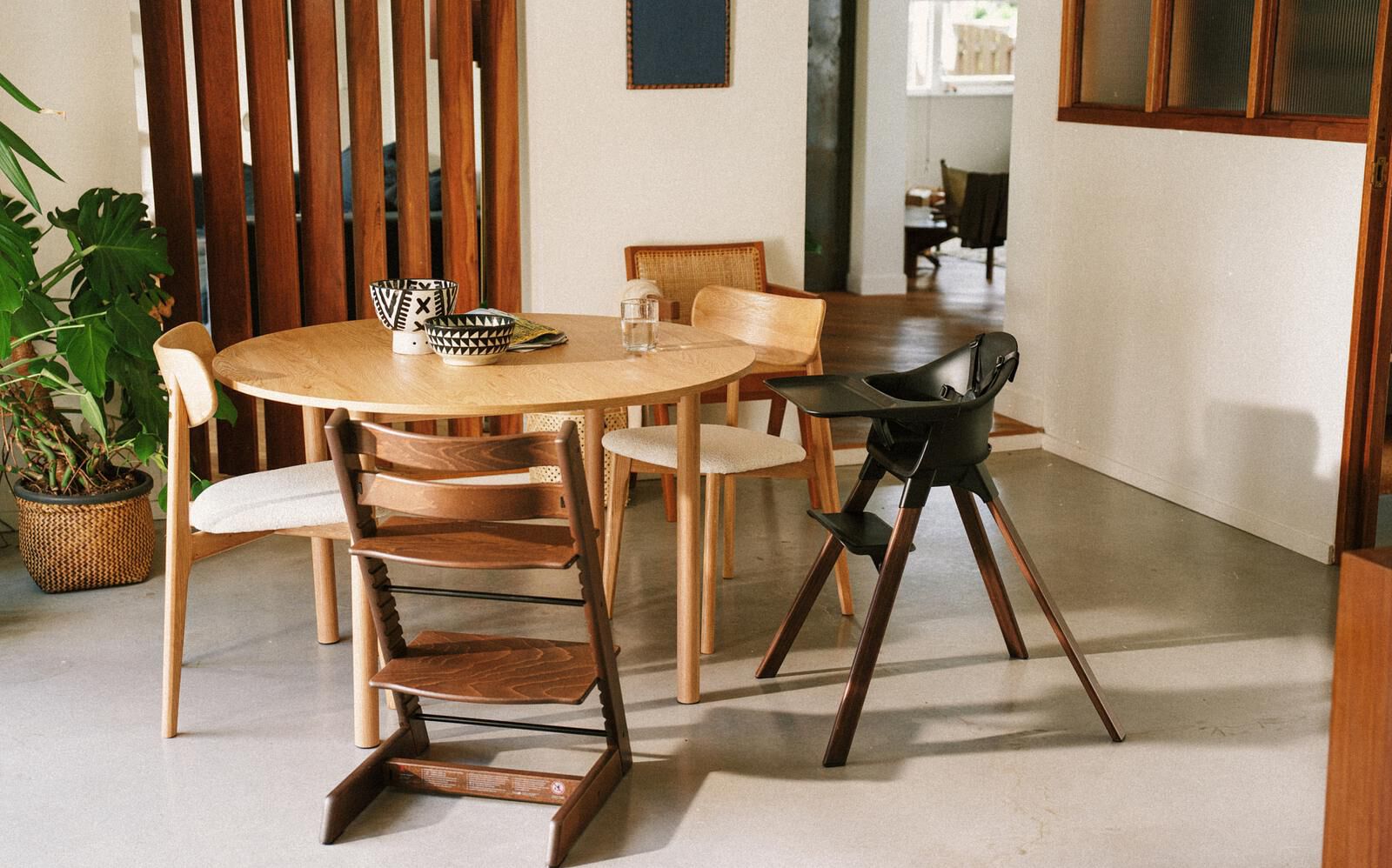A dining room with a round wooden table, chairs, a dark brown Tripp Trapp chair, and a black high chair.