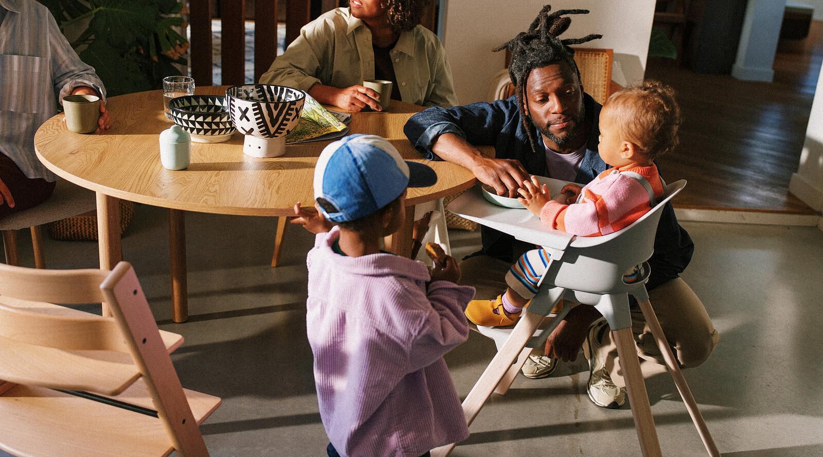 Family dinner: Man feeds baby in a high chair. Another child stands. A Tripp Trapp chair is visible.