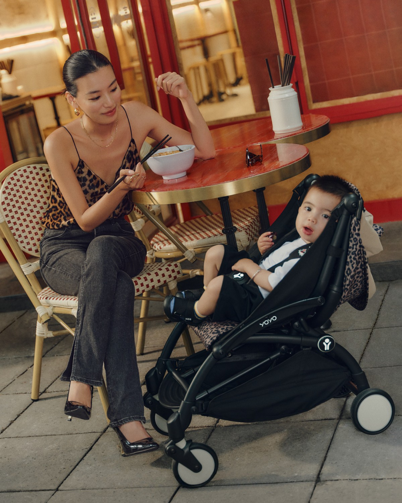 Vrouw eet aan cafétafel; baby kijkt toe vanuit een STOKKE YOYO-kinderwagen met luipaardprint.
