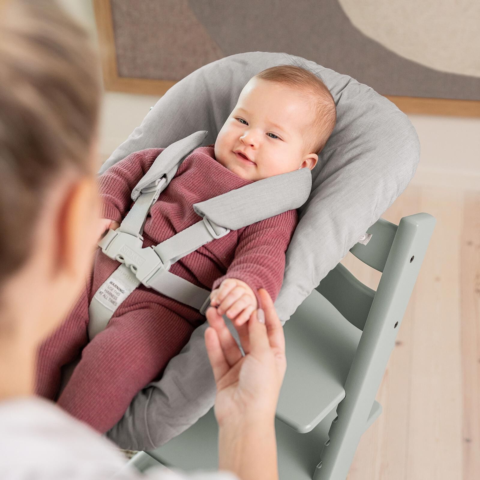 Smiling baby in a grey Tripp Trapp high chair with Newborn Set, holding an adult's hand.