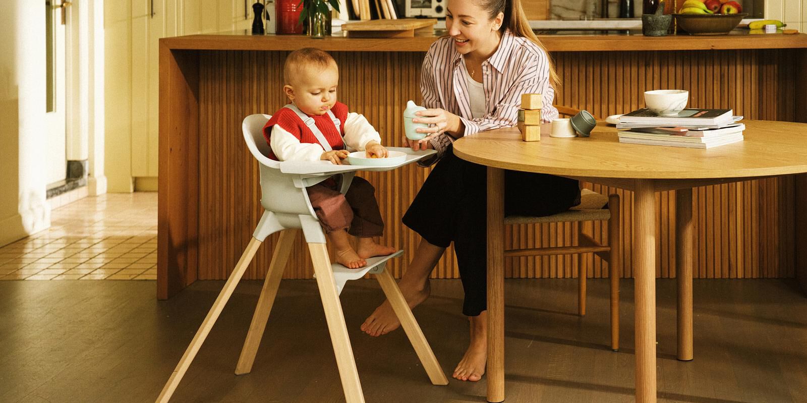 A baby eats in a light grey and wood high chair while a woman smiles beside them at a wooden table.