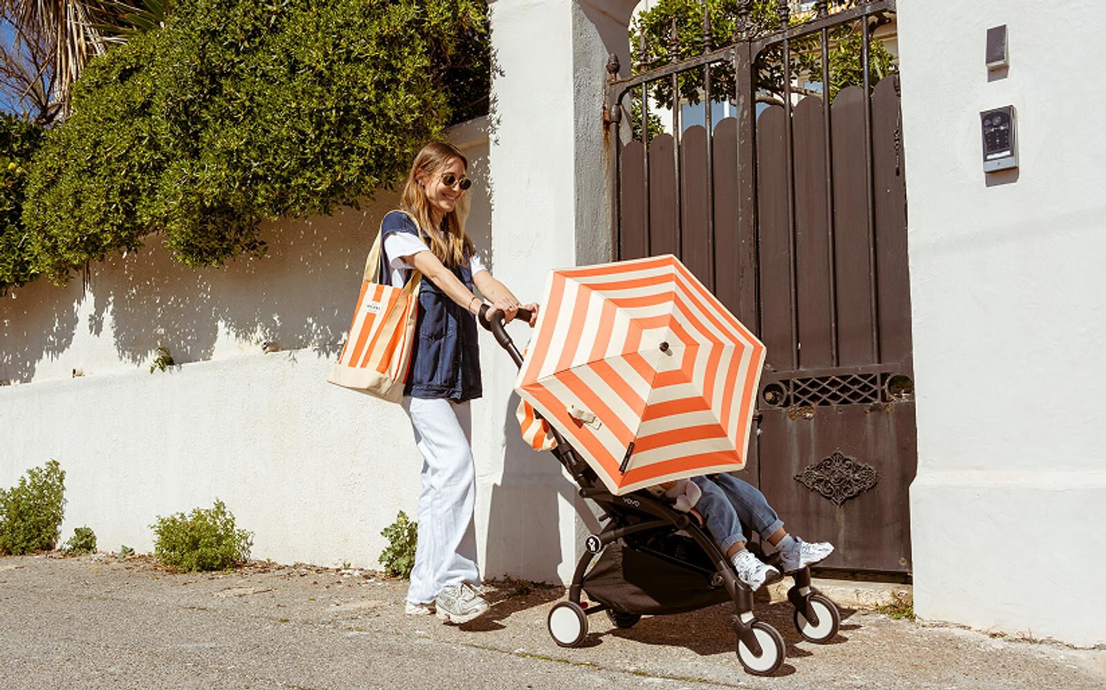 Frau schiebt einen STOKKE YOYO Kinderwagen mit einem orange gestreiften Regenschirm und Kind auf einer sonnigen Straße.