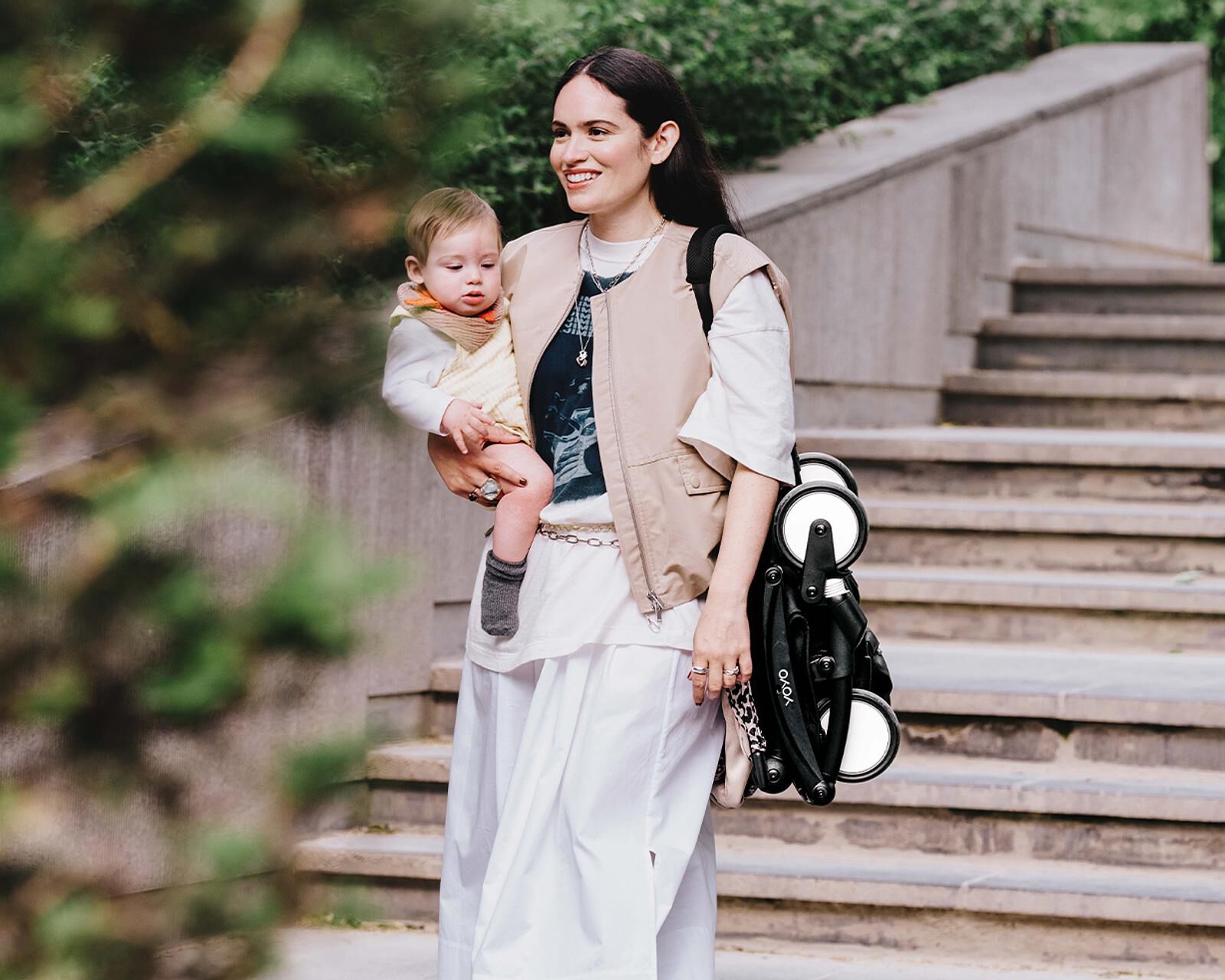 Eine Frau hält ein Baby und einen zusammengeklappten STOKKE YOYO Kinderwagen und lächelt draußen in der Nähe von Treppen.