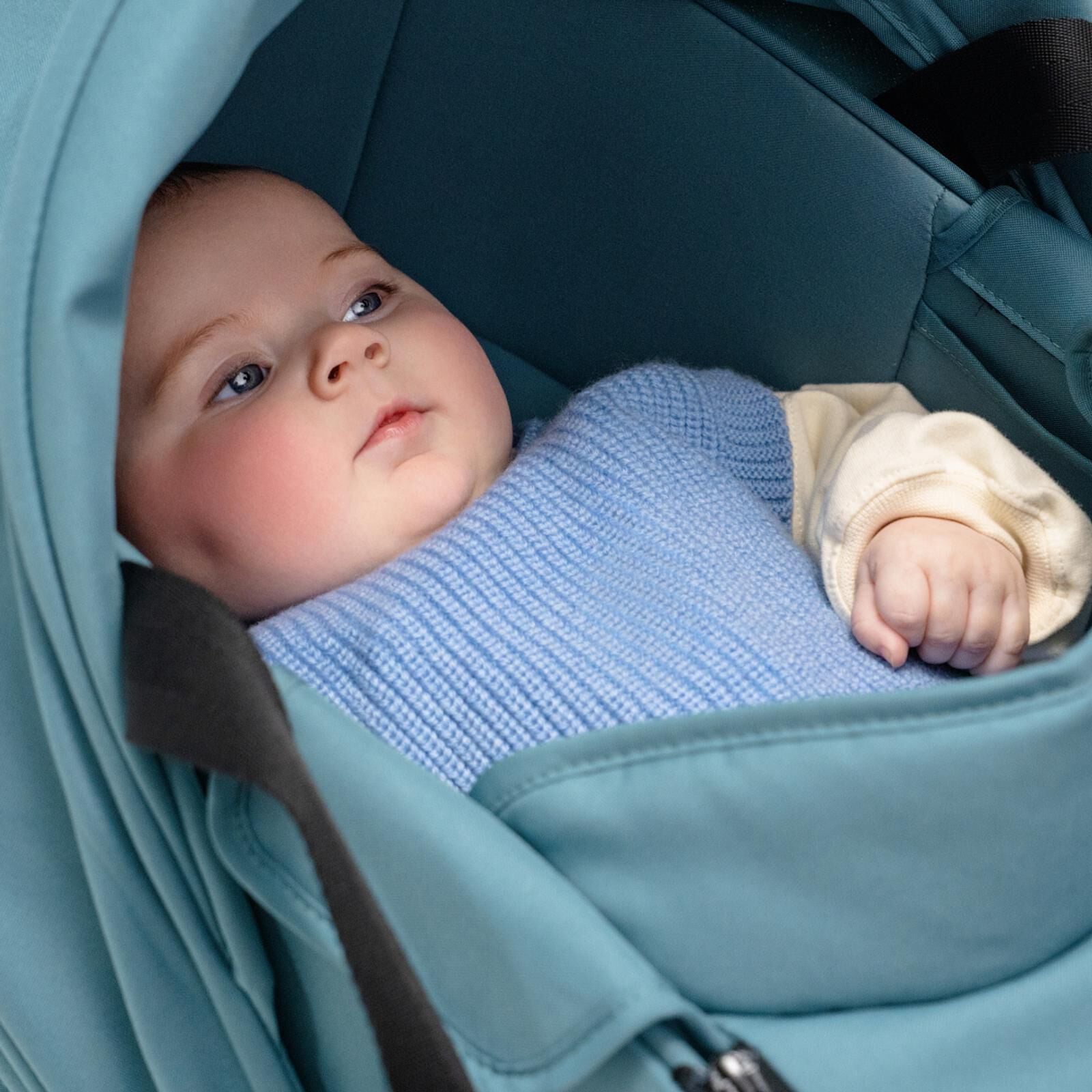 Ein Baby mit blauen Augen und rosigen Wangen liegt in einem türkisfarbenen STOKKE YOYO Kinderwagen und schaut nach oben.