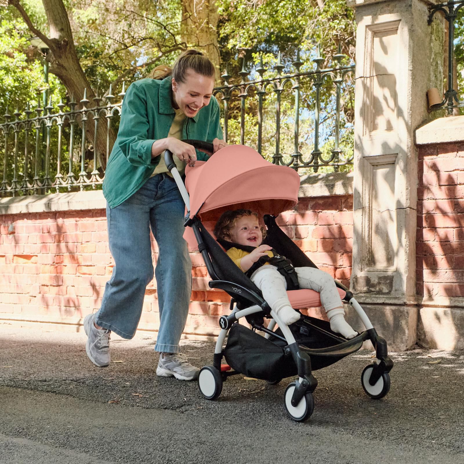 A smiling woman pushes a happy baby in a pink YOYO stroller on a sunny path.