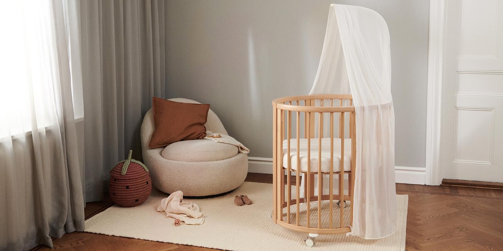 A cozy nursery room with a Stokke Sleepi Mini crib, a cream armchair, and a strawberry basket.