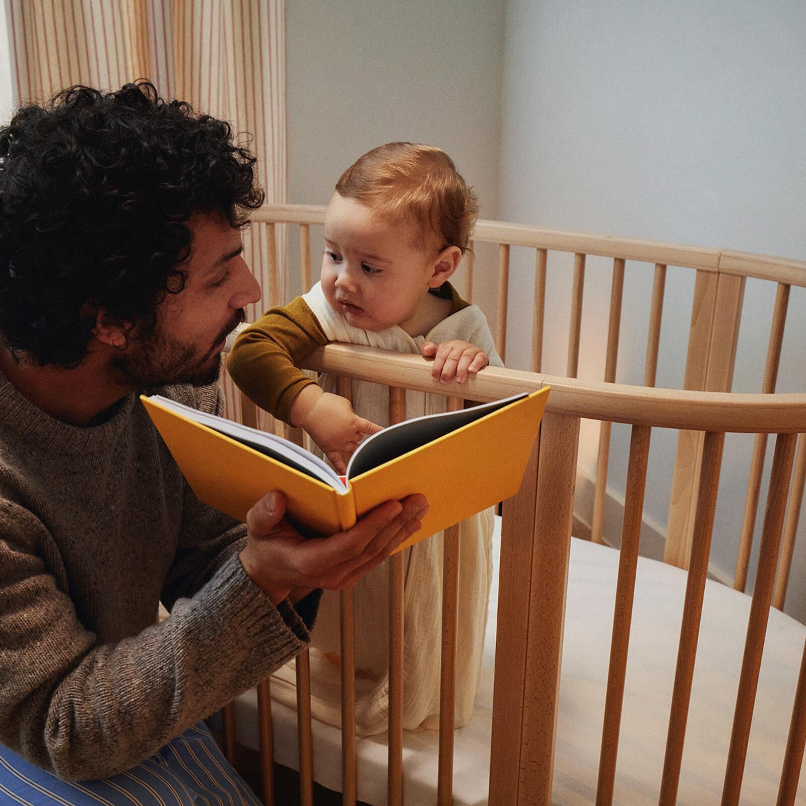 A father reads a story from a yellow book to his baby standing in a wooden crib.