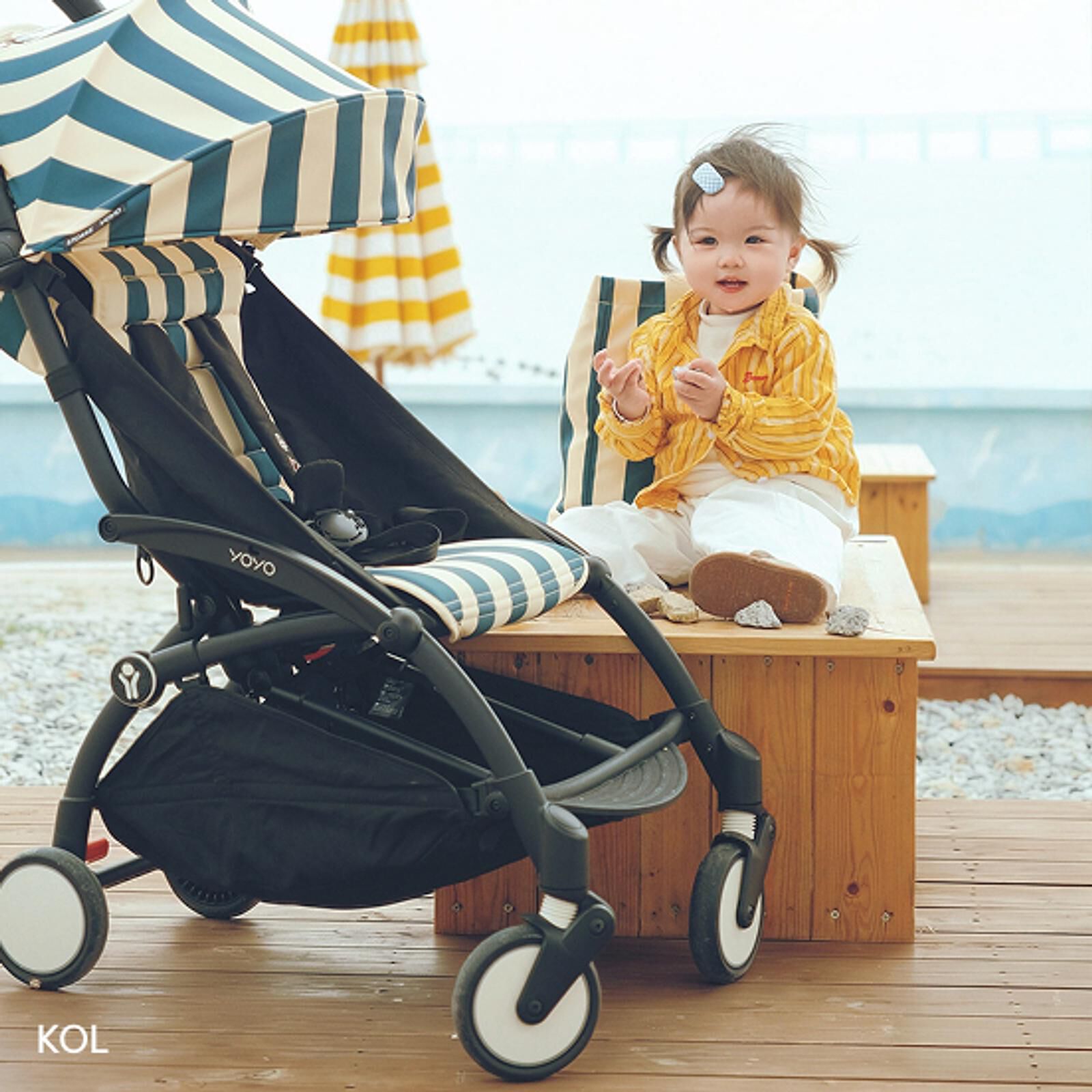 Ein lächelndes Kind, das Kieselsteine hält, sitzt auf einer Bank neben einem STOKKE YOYO Kinderwagen mit gestreiftem Verdeck.
