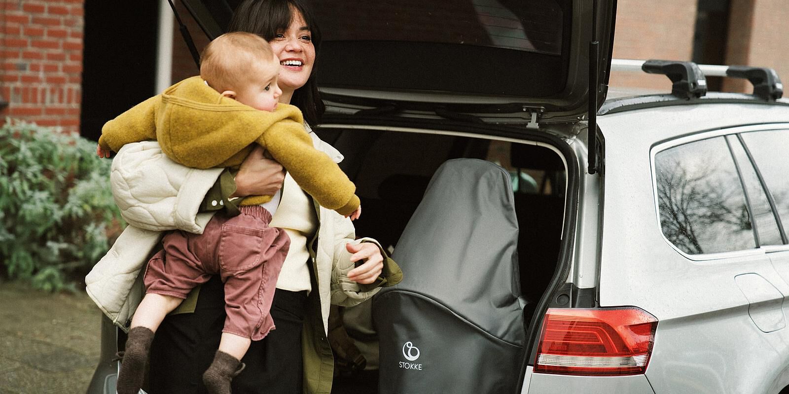 Mujer sonriente sostiene a un bebé, guardando el cochecito STOKKE YOYO en el maletero de un coche.