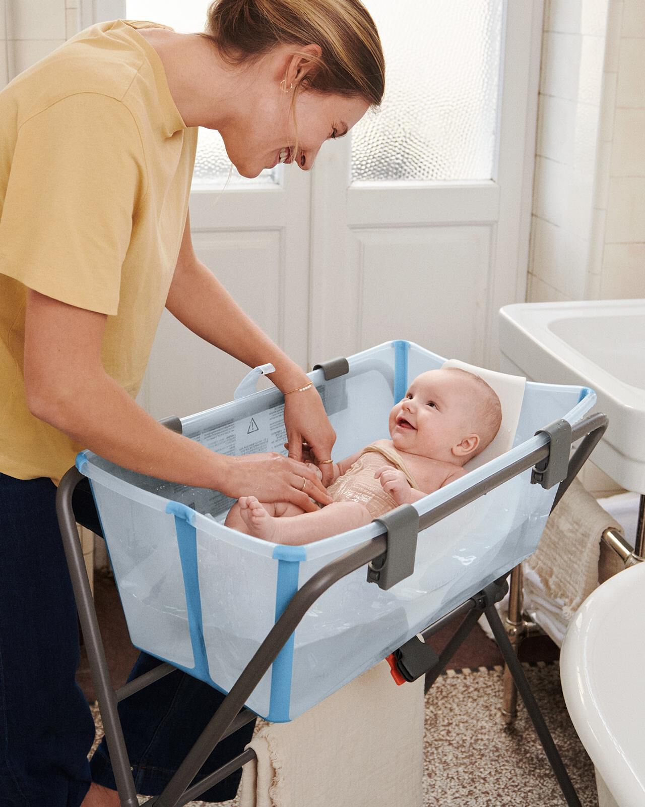 En kvinna badar en leende bebis i ett blått hopfällbart babybadkar på ett stativ.